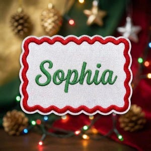 May include: A rectangular embroidered patch with a white center and a scalloped red border. The name Sophia is stitched in the center in a green cursive font. The patch features a textured fabric finish with clear, bold lettering and stitching.