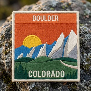 May include: A square embroidered patch featuring a landscape with the text BOULDER at the top and COLORADO at the bottom. The design shows a yellow sun, blue mountains, white rocky peaks, and green hills with a path and pine trees.