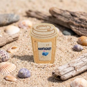 May include: A tan embroidered patch shaped like a disposable coffee cup with a lid. The cup features the text NANTUCKET in dark blue letters above a small blue whale icon with a tiny pink heart, all rendered in detailed thread stitching.