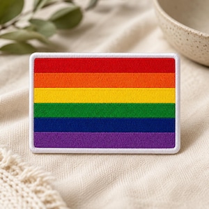 May include: Rectangular patch featuring the rainbow flag design. The patch has a white border and is embroidered with horizontal stripes in red, orange, yellow, green, blue, and purple. The patch is likely for clothing or accessories.