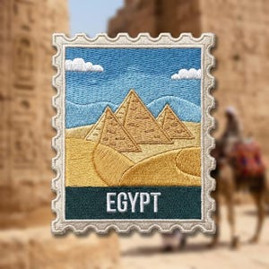 May include: A rectangular embroidered patch shaped like a postage stamp featuring a desert landscape with three golden pyramids under a blue sky with white clouds. The word EGYPT is stitched in white capital letters on a dark teal background.