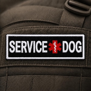 May include: A rectangular black patch with a white border featuring the white text SERVICE DOG and a red Star of Life symbol with a white serpent and staff emblem in the center, attached to a dark olive green fabric surface with webbing.