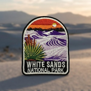 May include: An embroidered patch featuring a landscape of White Sands National Park with a green yucca plant in the foreground, purple sand dunes, and an orange sunset sky with a white sun. The text reads WHITE SANDS NATIONAL PARK in white.