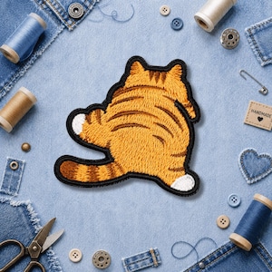 May include: Embroidered orange tabby cat patch with brown stripes and a black border. The cat patch has a white paw and is surrounded by sewing supplies, including denim scraps, thread spools, and buttons.