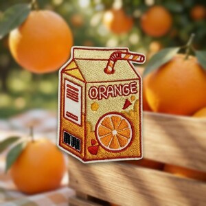 May include: An embroidered patch shaped like a carton of orange juice. The patch is yellow and orange with the word ORANGE printed in red letters. It features a graphic of a sliced orange and a red and white striped straw sticking out the top.