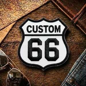 May include: A shield-shaped embroidered patch featuring a white background with a thick black border. The text CUSTOM is stitched in black at the top, with the large black numbers 66 centered below, mimicking the classic Route 66 road sign design.