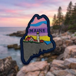 May include: An embroidered patch shaped like the state of Maine with a dark blue border. The design features a lighthouse and building against a sunset sky with pink, purple, and yellow hues, with the word MAINE printed in dark letters.