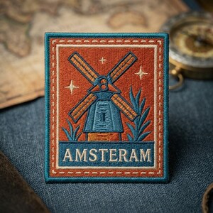 May include: A rectangular embroidered patch featuring a central blue windmill on an orange background, surrounded by small stars and foliage. The word AMSTERAM is stitched in white capital letters within a blue horizontal band at the bottom.