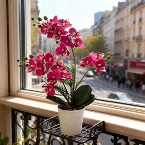 May include: A potted artificial orchid plant with vibrant magenta flowers and dark green leaves. The plant is housed in a white, textured cylindrical pot, which sits on a black metal balcony railing shelf against a bright window background.