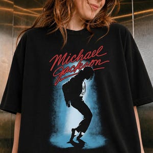 May include: A black short-sleeved t-shirt featuring a graphic print of a silhouette of Michael Jackson in a dance pose. The design includes the name Michael Jackson in red cursive script above the figure, set against a blue spotlight effect.