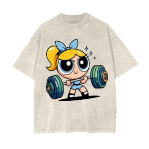May include: Beige t-shirt featuring a cartoon character lifting weights. The character has blonde hair, a blue bow, and is wearing a blue outfit. The barbell has green and blue weights. The design is centered on the shirt.