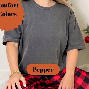 Pode incluir: Uma camiseta cinza escuro com a palavra "Pepper" em um oval marrom. A pessoa est&aacute; usando cal&ccedil;as de pijama xadrez vermelhas e pretas. A camisa faz parte da cole&ccedil;&atilde;o Comfort Colors.