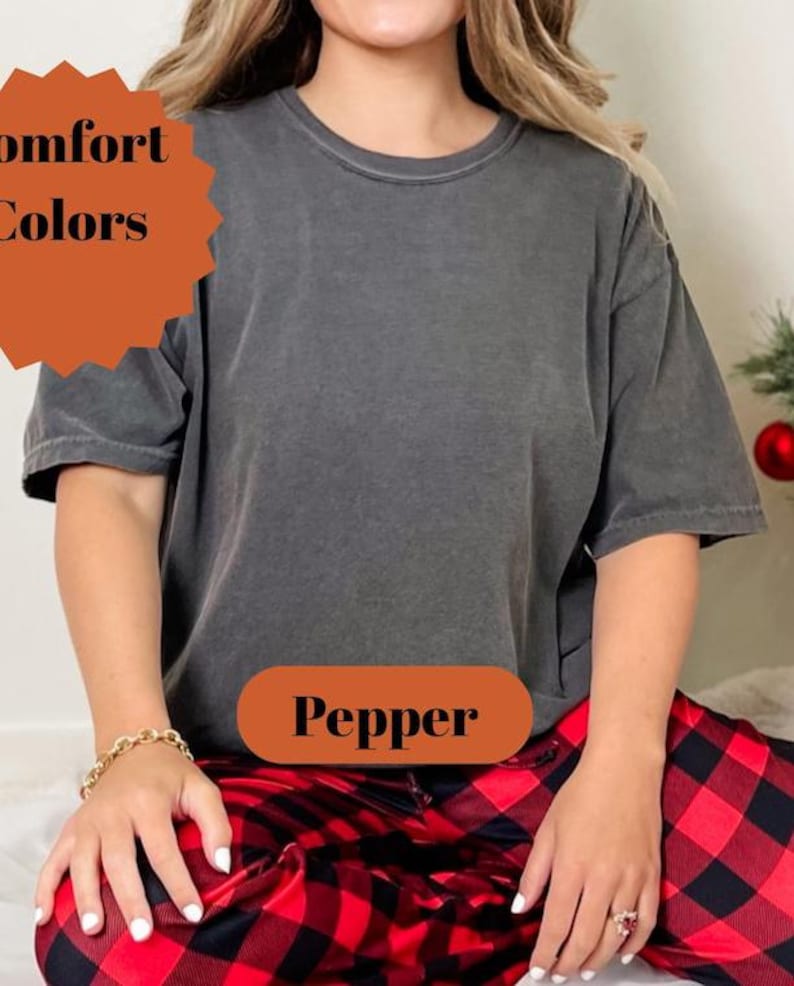 Pode incluir: Uma camiseta cinza escuro com a palavra "Pepper" em um oval marrom. A pessoa est&aacute; usando cal&ccedil;as de pijama xadrez vermelhas e pretas. A camisa faz parte da cole&ccedil;&atilde;o Comfort Colors.