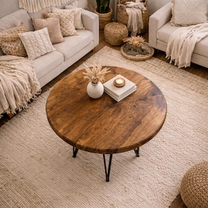May include: A round, wooden coffee table with black metal legs is the focal point of a cozy living room. The table is adorned with a white vase, dried flowers, a stack of books, and a lit candle. Beige sofas, pillows, and a rug complete the warm, inviting space.