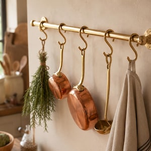 Pode incluir: Uma barra de cozinha de latão montada na parede com cinco ganchos em S a condizer. Pendurados nos ganchos estão um molho de ervas frescas, dois tachos de cobre polido com pegas de latão, uma concha de latão e um pano de cozinha de linho bege.