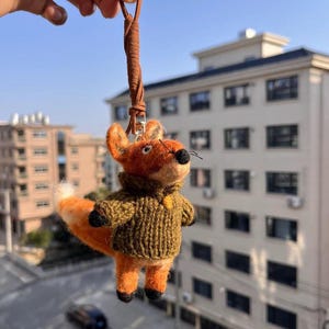 Puede incluir: Un llavero de zorro naranja hecho a mano con un suéter marrón y una correa de cuero. El zorro tiene una cola con punta blanca, patas negras y una nariz negra. El llavero está suspendido frente a un edificio.
