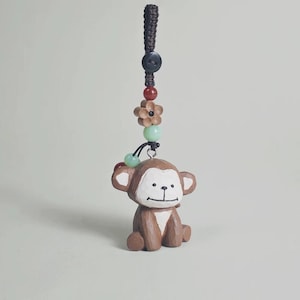 Puede incluir: Una figura de mono de madera marrón con detalles blancos, una cara sonriente y un cordón decorativo. El cordón incluye un botón negro, cuentas rojas y verdes y una flor de madera. Un artículo encantador y artesanal.