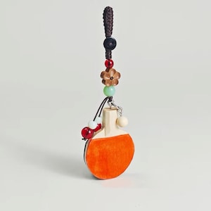 May include: A decorative ping pong paddle keychain with an orange paddle and a wooden handle. The keychain features a brown braided cord, a black bead, a flower-shaped bead, and various colorful beads.