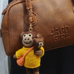 May include: A brown leather handbag with the embossed text "JAGD GIRL" is shown. A small, handmade monkey keyring with a yellow sweater and orange bag hangs from the handle. The monkey is holding a smaller bear.