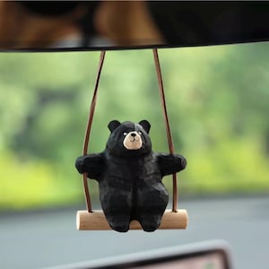 May include: A black wooden bear figurine on a swing. The bear has a light-coloured face and is suspended by brown cords from a wooden bar. The background is a blurred green and grey.