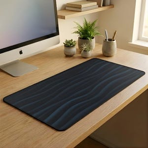 Op de afbeelding: Een rechthoekige bureaumat met een donkerblauw golvend patroon. De mat ligt op een licht houten bureau, met een computermonitor, potplanten en bureau-accessoires op de achtergrond. De mat is ontworpen om het bureaublad te beschermen.
