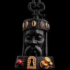 May include: A black sculpture of a face wearing a crown with gold accents. The crown features oval gold details. Below the face, the base displays a red faceted gem, a gold keyhole plate, and a cluster of gold coins against a black background.