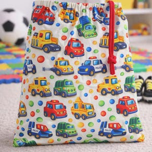 Puede incluir: Una bolsa blanca con cordón con un patrón colorido de coches de juguete, camiones y vehículos de policía en rojo, amarillo, verde y azul. La bolsa tiene un cierre de cordón rojo y está decorada con lunares de colores.
