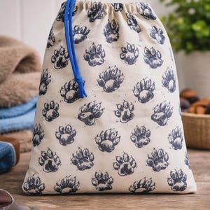 Puede incluir: Una bolsa con cordón de color crema con un patrón repetido de huellas de patas azul oscuro. La bolsa tiene un cierre de cordón azul. La bolsa está sobre una superficie de madera.