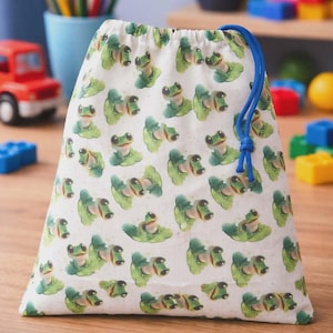 Puede incluir: Una bolsa blanca con cordón con un patrón repetido de ranas verdes sobre nenúfares. La bolsa tiene un cierre de cordón azul. El fondo incluye juguetes y lápices de colores.