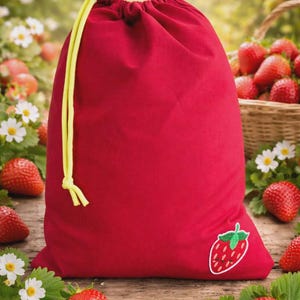 Puede incluir: Una bolsa roja con cordón y cierre de cordón amarillo y un parche de fresa. Fresas frescas y margaritas están esparcidas sobre una superficie de madera. Una cesta de fresas está en el fondo.