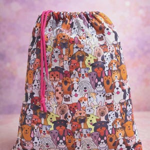 Puede incluir: Una bolsa con cordón con un patrón colorido de varias razas de perros. La bolsa tiene un cierre de cordón rosa. La tela presenta un patrón repetido de ilustraciones de perros en tonos marrones, blancos y negros.