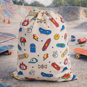 Puede incluir: Una bolsa con cordón de color crema con un patrón repetido de patinetas, rayos, llamas y otros gráficos relacionados con el skateboarding en rojo, azul, amarillo y negro. La bolsa está sobre una superficie de hormigón con un parque de patinaje en el fondo.