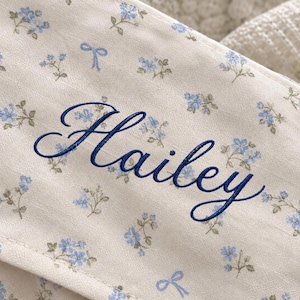 May include: A cream-colored fabric featuring a repeating pattern of small blue flowers and light blue bows. The name Hailey is embroidered in dark blue cursive script across the center of the fabric, which has a visible stitched seam.