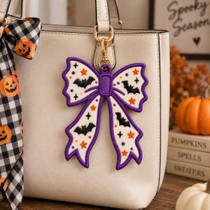 Peut inclure: Un sac à main blanc avec un grand bijou de sac brodé en forme de nœud. L'ornement présente une bordure violette et est décoré de silhouettes de chauves-souris noires, d'étoiles orange et de petites étoiles noires. Il s'attache avec un fermoir doré.