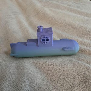 Puede incluir: Un submarino de juguete impreso en 3D en tonos azules y verdes. El submarino tiene una pequeña cabina con una ventana circular y una chimenea. El juguete está sobre una superficie beige.