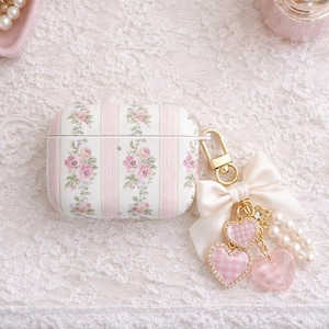 May include: A white and pink floral patterned AirPod case with a gold-colored clasp and a charm. The charm includes a white bow, two pink heart charms, and a string of pearls. The case has pink stripes.