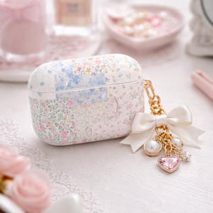 May include: A white and floral patterned case for earbuds. The case has a gold chain with a white bow, a pink heart charm, and pearl accents. The case is decorated with a patchwork of floral patterns in pink, blue, and white.