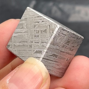 May include: A metallic cube featuring a distinct Widmanstätten pattern on its surface. The object displays a silver-gray color with a complex, intersecting grid of lines and crystalline textures across its visible faces, showcasing a geometric form.