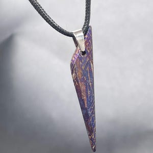 May include: A triangular pendant featuring a metallic surface with a purple and copper-toned pattern, suspended from a black cord by a silver-colored metal bail. The pendant has a sharp, pointed bottom edge and a textured, geometric appearance.