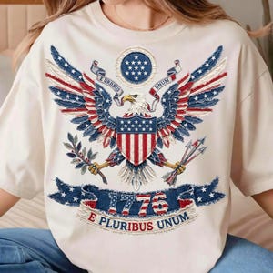 May include: Cream-colored t-shirt featuring a patriotic design. The design includes a detailed eagle with outstretched wings, incorporating red, white, and blue elements reminiscent of the American flag. The year "1776" is prominently displayed.