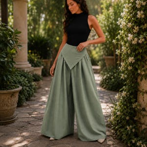 Könnte beinhalten: Ein schwarzes ärmelloses Oberteil kombiniert mit einer hoch taillierten, weit geschnittenen Hose in einem weichen Salbeigrün. Die Hose hat ein einzigartiges, gefaltetes Design an der Taille. Das Outfit wird im Freien getragen, umgeben von Grün und Steinarchitektur.