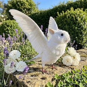 May include: A whimsical white rabbit sculpture with feathered wings, standing on a stone wall. The rabbit has black eyes and is surrounded by white roses and lavender. The wings are spread, giving the impression of flight.