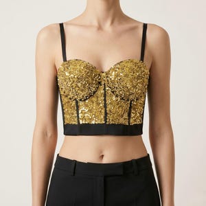 May include: A gold sequined bustier top with black trim and straps. The top features a sweetheart neckline and a fitted silhouette. The garment is worn with black pants.