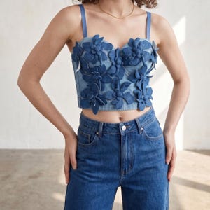 May include: A denim crop top with floral embellishments. The top features a sweetheart neckline and thin straps. The flowers are a darker shade of blue than the top. The model is also wearing blue jeans.