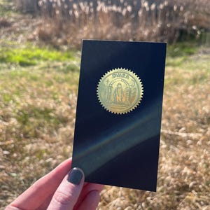 May include: A black rectangular card with a gold seal featuring the word "Dvara" and a figure. The card is held against a blurred outdoor background, with sunlight creating a rainbow effect.