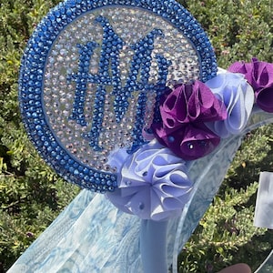 May include: A headband featuring a circular ear covered in blue and clear rhinestones with a dark blue monogram of the letters HA in the center. The headband is decorated with purple and light purple fabric rosettes and a light blue veil.
