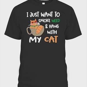 May include: A black short-sleeve t-shirt featuring a graphic of an orange cat sitting in a mug, wearing sunglasses with marijuana leaf patterns, and smoking a cigarette. The text reads: I JUST WANT TO SMOKE WEED & HANG WITH MY CAT.