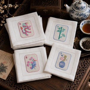 May include: Set of four white linen cocktail napkins with embroidered designs. Each napkin features a different image: a dragon, bamboo, a bird with flowers, and a vase of flowers. The napkins are arranged on a dark wooden surface.