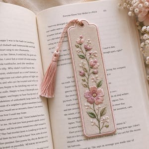 May include: A rectangular fabric bookmark with rounded corners, featuring intricate embroidery of pink flowers and green leaves on a beige background. The bookmark includes a matching pink tassel attached to a metal eyelet at the top edge.