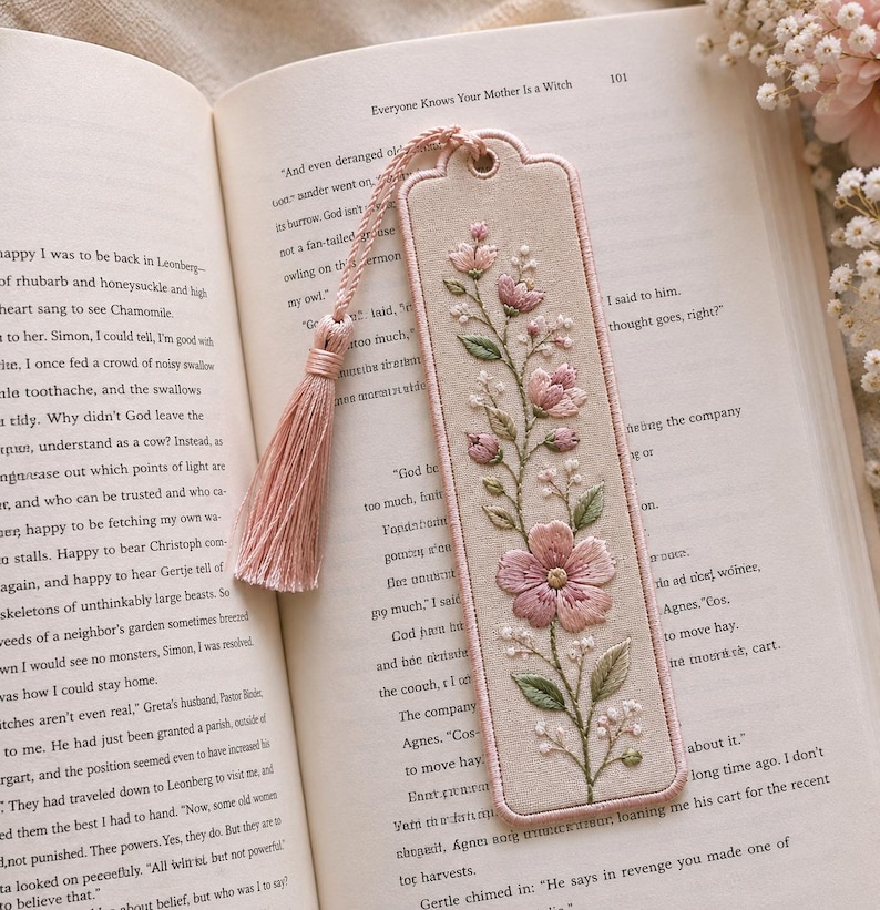 May include: A rectangular fabric bookmark with rounded corners, featuring intricate embroidery of pink flowers and green leaves on a beige background. The bookmark includes a matching pink tassel attached to a metal eyelet at the top edge.